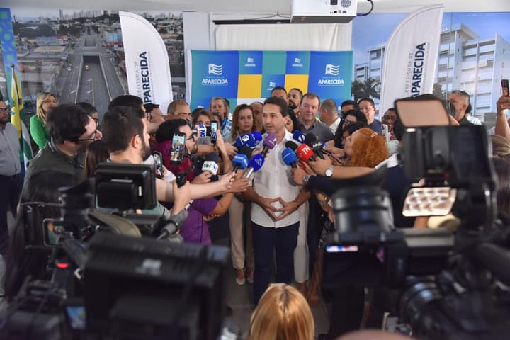 Imagem de jornalistas em pé, com câmeras e microfones, apontados para o prefeito